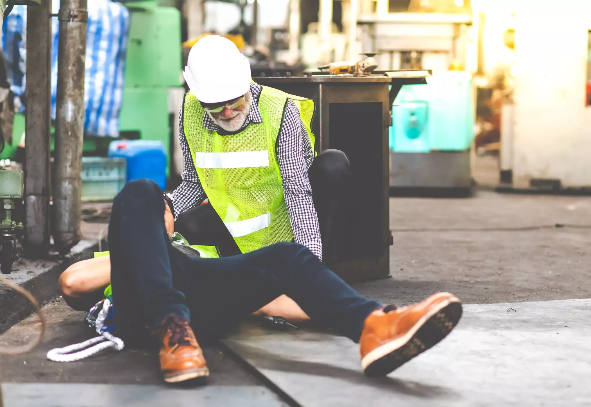 An injured man laying on the ground with a co-worker who is wearing a hard hat and a neon safety vest.