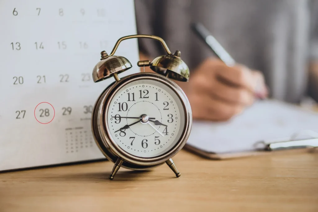 A calendar and a clock sitting on a table, while a person writes in background.