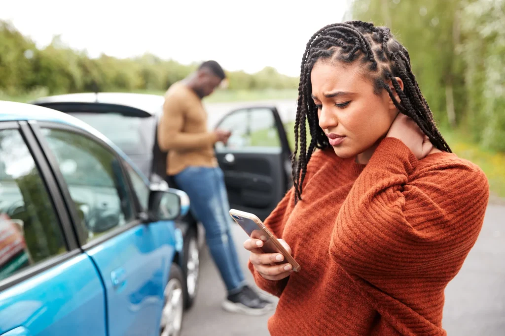 A woman holding her neck and using her phone after a car accident, with another driver in the background near their vehicles, representing the process of documenting and reporting a crash in San Antonio.